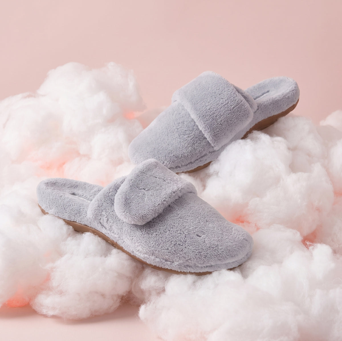 A pair of plush light gray slip-on slippers with a wide strap, styled on a bed of fluffy white faux clouds against a soft pink background.