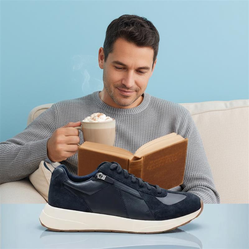 Man reading a book and holding a hot beverage with a navy blue sneaker in the foreground.