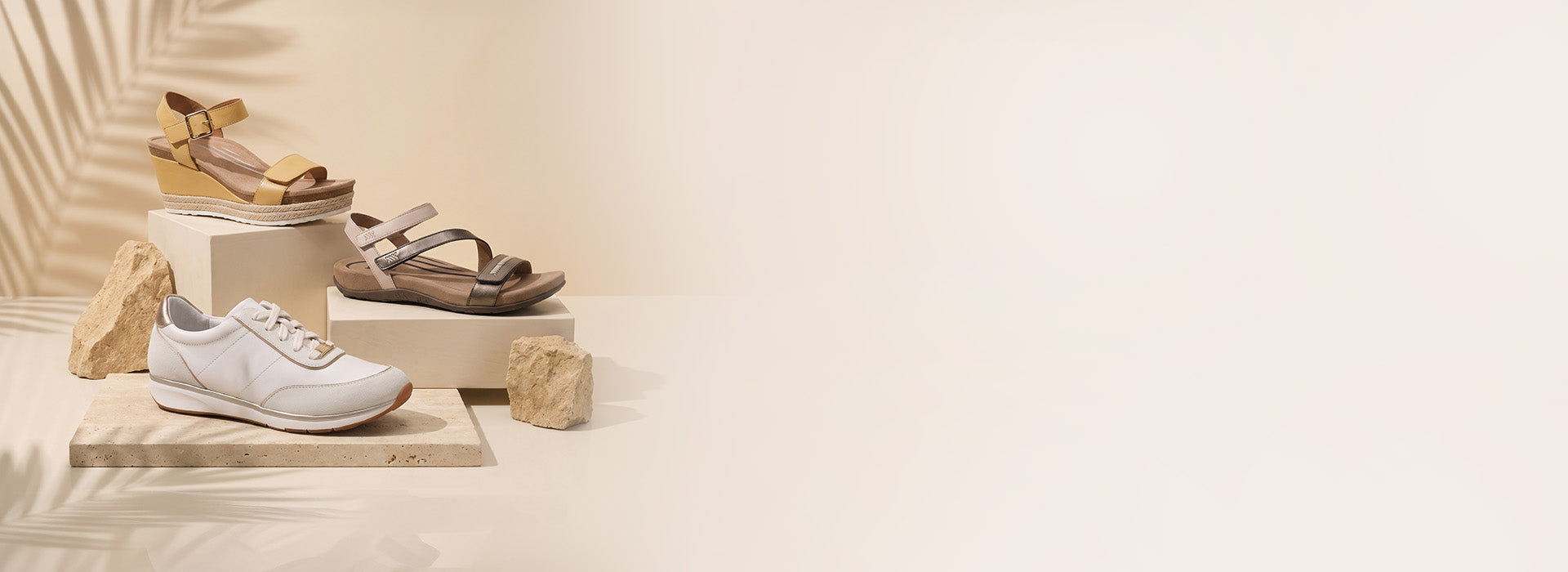 A styled product display featuring a white low-top sneaker, a brown strappy sandal, and a tan wedge sandal arranged on stacked beige blocks with small stone props against a warm cream background.