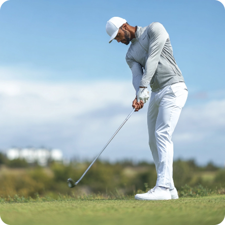 Golfer in a gray shirt and white pants on a golf course with a clear blue sky.
