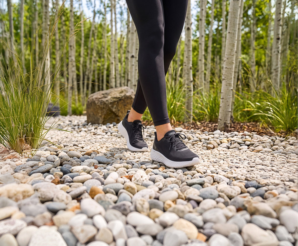 A person wearing black athletic leggings and black sneakers with white soles walking on a gravel path in a wooded outdoor setting.
