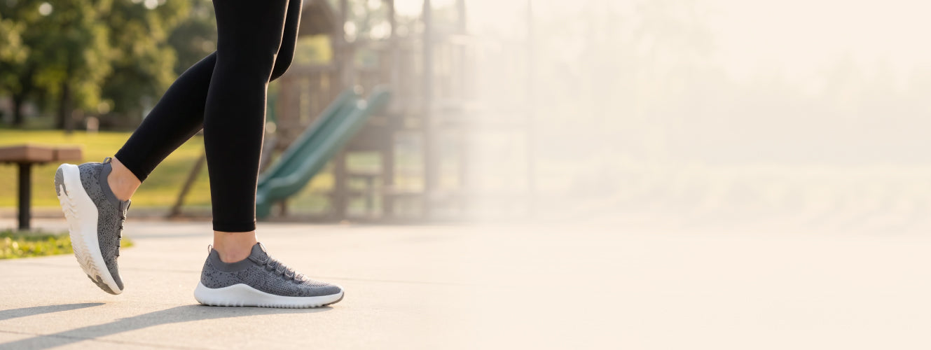 Person wearing gray sneakers and black leggings walking on a path with a playground in the background.