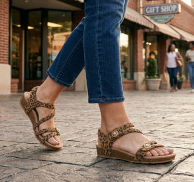 A person wearing leopard-print flat sandals with blue jeans walking on a cobblestone street in front of a brick storefront.