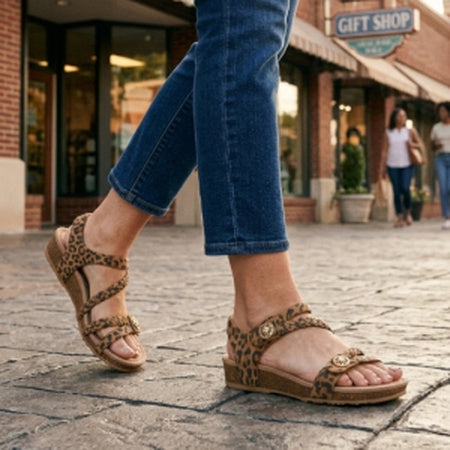 A person wearing leopard-print flat sandals with blue jeans walking on a cobblestone street in front of a brick storefront.