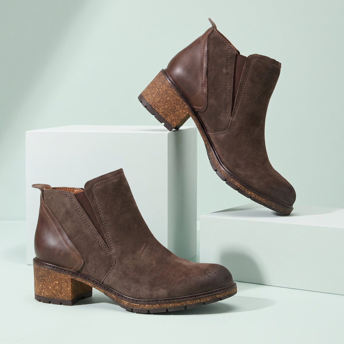 A pair of dark brown suede Chelsea-style ankle boots with chunky cork block heels, displayed on white geometric pedestals against a pale mint green background.