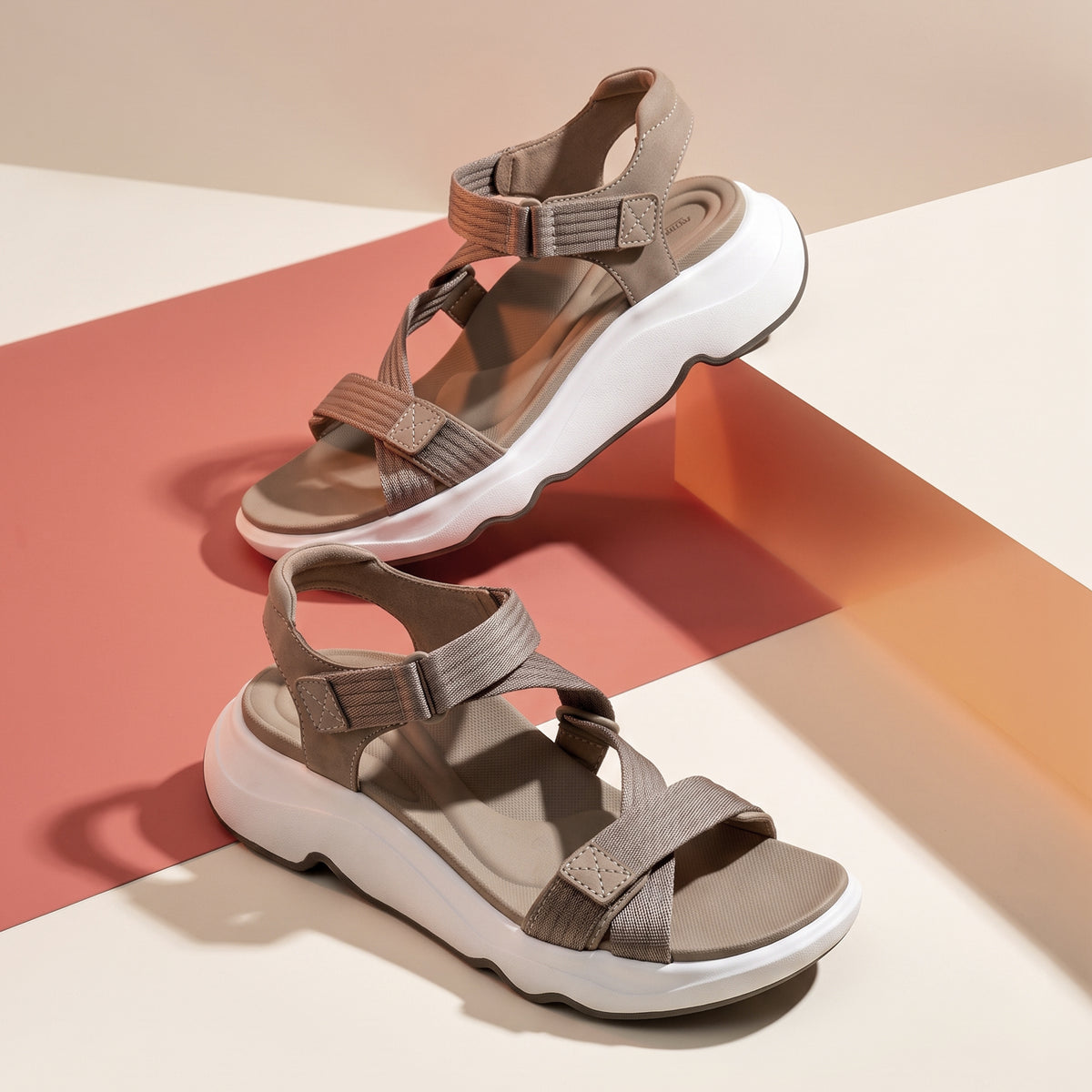 A pair of chunky platform sandals in taupe and white, styled on a geometric studio background with pink and orange panels.