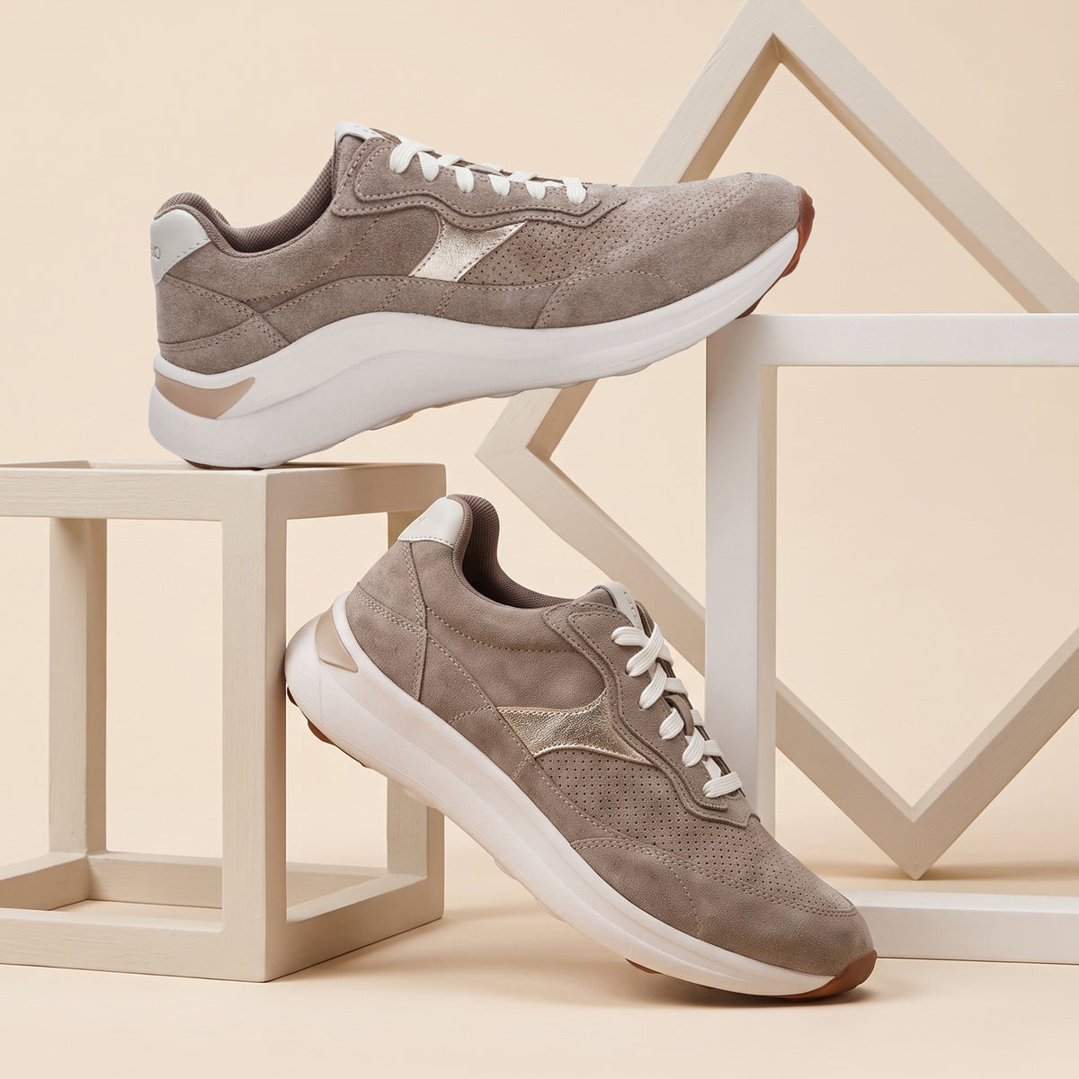 A pair of taupe-brown suede sneakers with white laces and white midsoles, styled on geometric beige display blocks against a warm beige background.