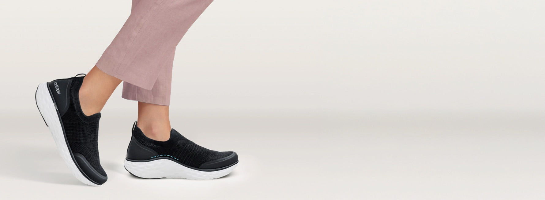Black slip-on shoes worn with light pink pants on a white background