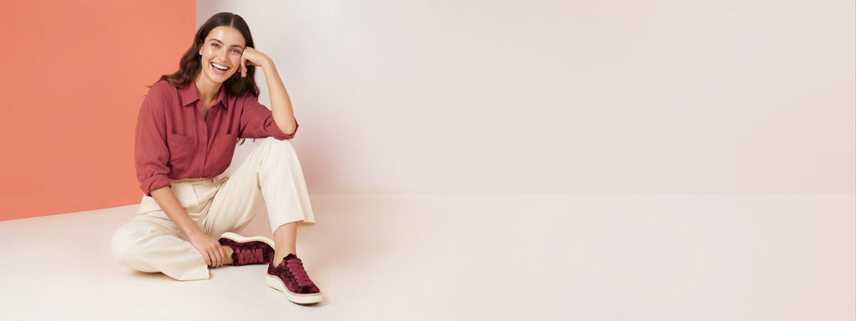 A woman sitting on the floor in a studio setting, wearing a dusty rose button-up shirt, cream wide-leg trousers, and burgundy low-top sneakers with white soles. The background is split diagonally between a warm peach-pink color and a light off-white color.