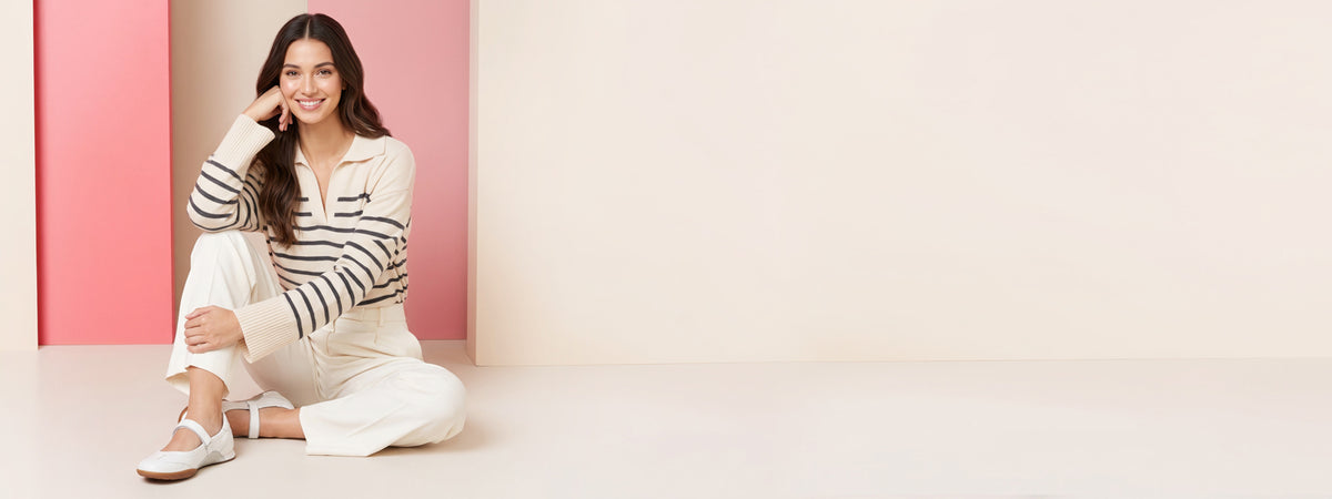A woman sits cross-legged on a light beige floor against a pink and beige wall, wearing a cream and navy striped long-sleeve polo shirt with white cropped pants and white sneakers.