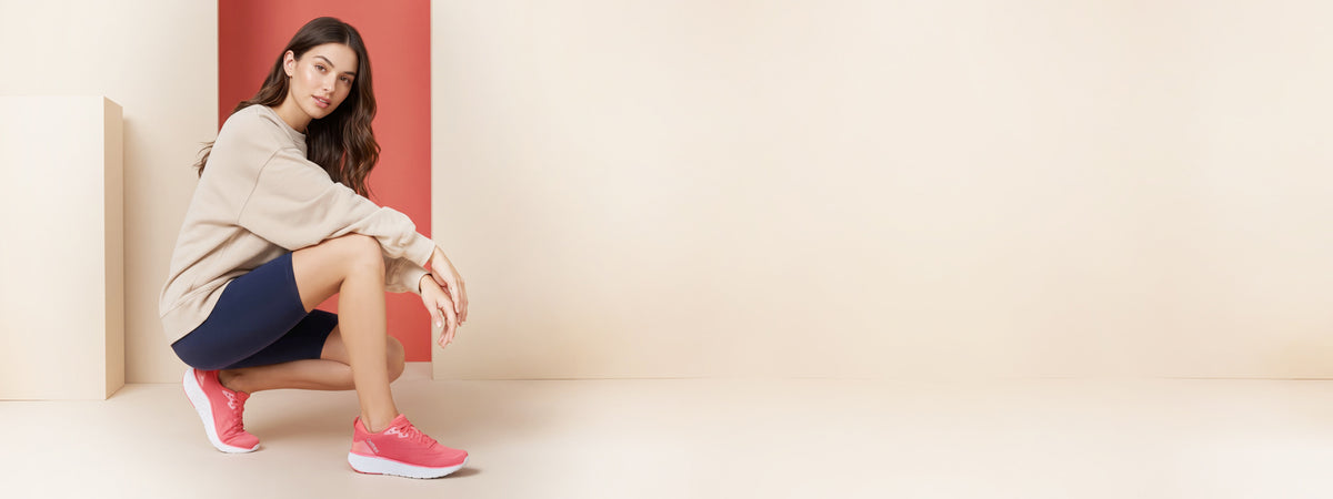 A woman crouches in a studio setting wearing a beige oversized sweatshirt, navy athletic shorts, and bright pink sneakers against a minimalist beige and coral background.