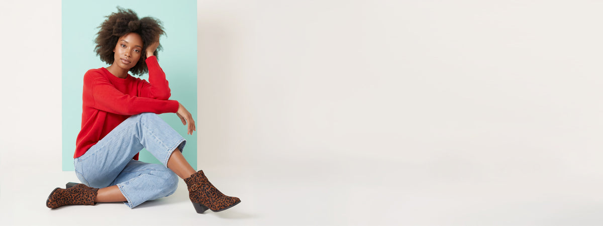 A woman with curly dark hair sits on the floor wearing a bright red long-sleeve top, light blue cropped jeans, and brown leopard-print ankle boots against a light blue and white studio background.