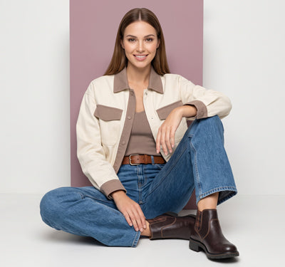 A woman sits cross-legged wearing a cream and taupe utility-style jacket, blue jeans, a brown top, and dark brown ankle boots against a studio backdrop with a mauve vertical panel.