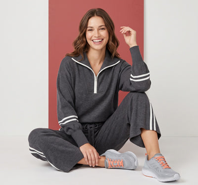 A woman sits cross-legged wearing a dark gray matching lounge set with white stripe accents on the sleeves and pants, paired with gray sneakers, against a studio backdrop with a muted red rectangular panel.