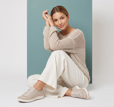 A woman sits on the floor wearing a chunky beige knit sweater, white wide-leg pants, and beige perforated sneakers, posed against a minimalist studio backdrop with a vertical teal panel.