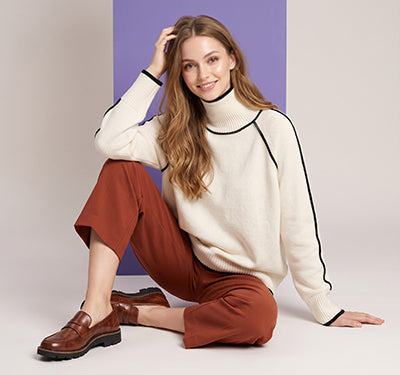 A woman sits cross-legged wearing a cream turtleneck sweater with black trim, rust-brown cropped pants, and brown loafers, posed against a studio backdrop with a purple panel.