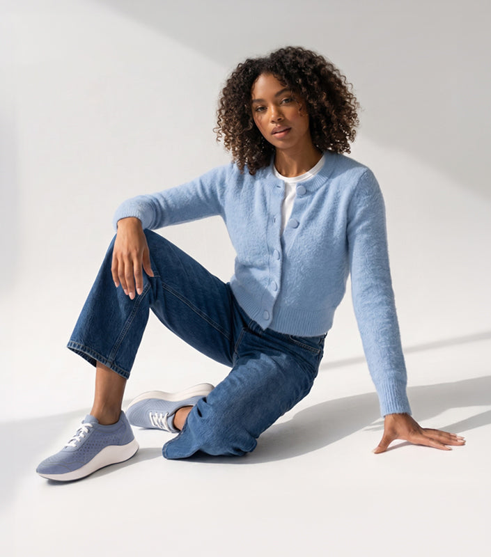 A woman with curly hair sits on a white studio floor wearing a light blue knit cardigan over a white top, blue jeans, and light blue sneakers.