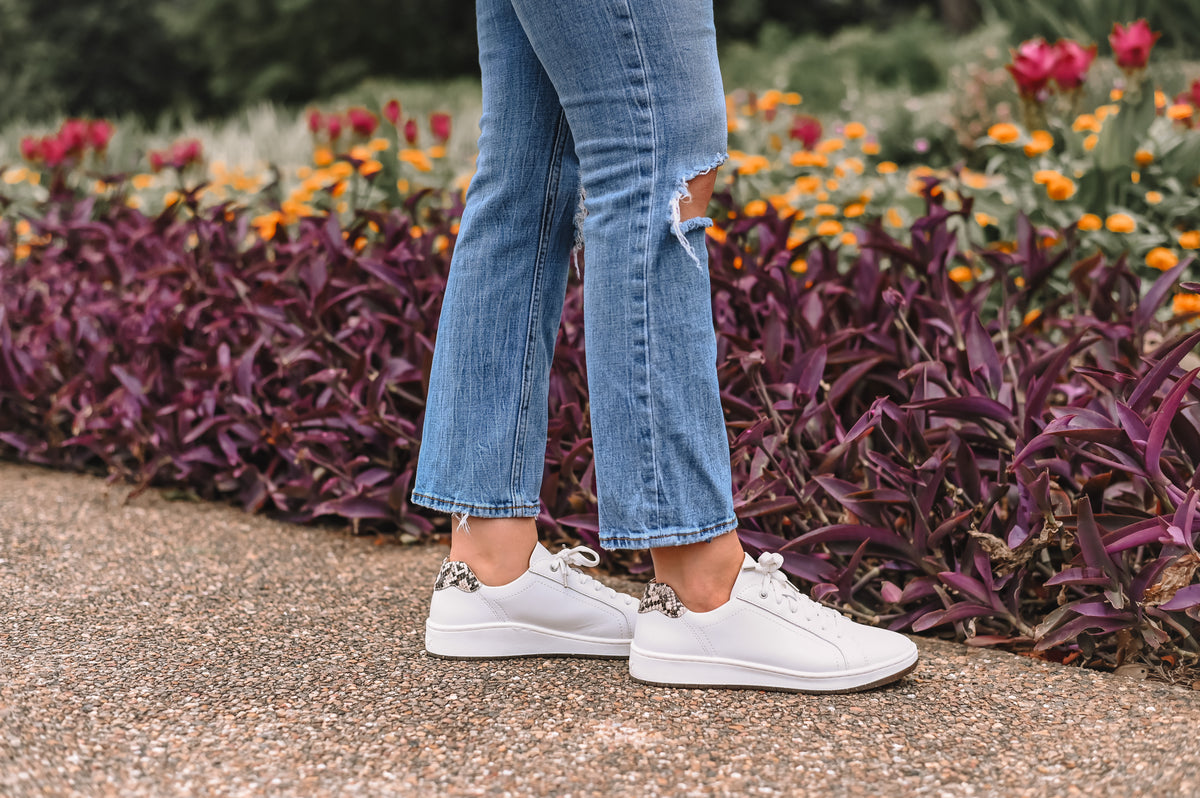 White leather sneakers with silver chain details on the laces, worn by a person in ripped blue jeans standing on a gravel path with a garden background.
