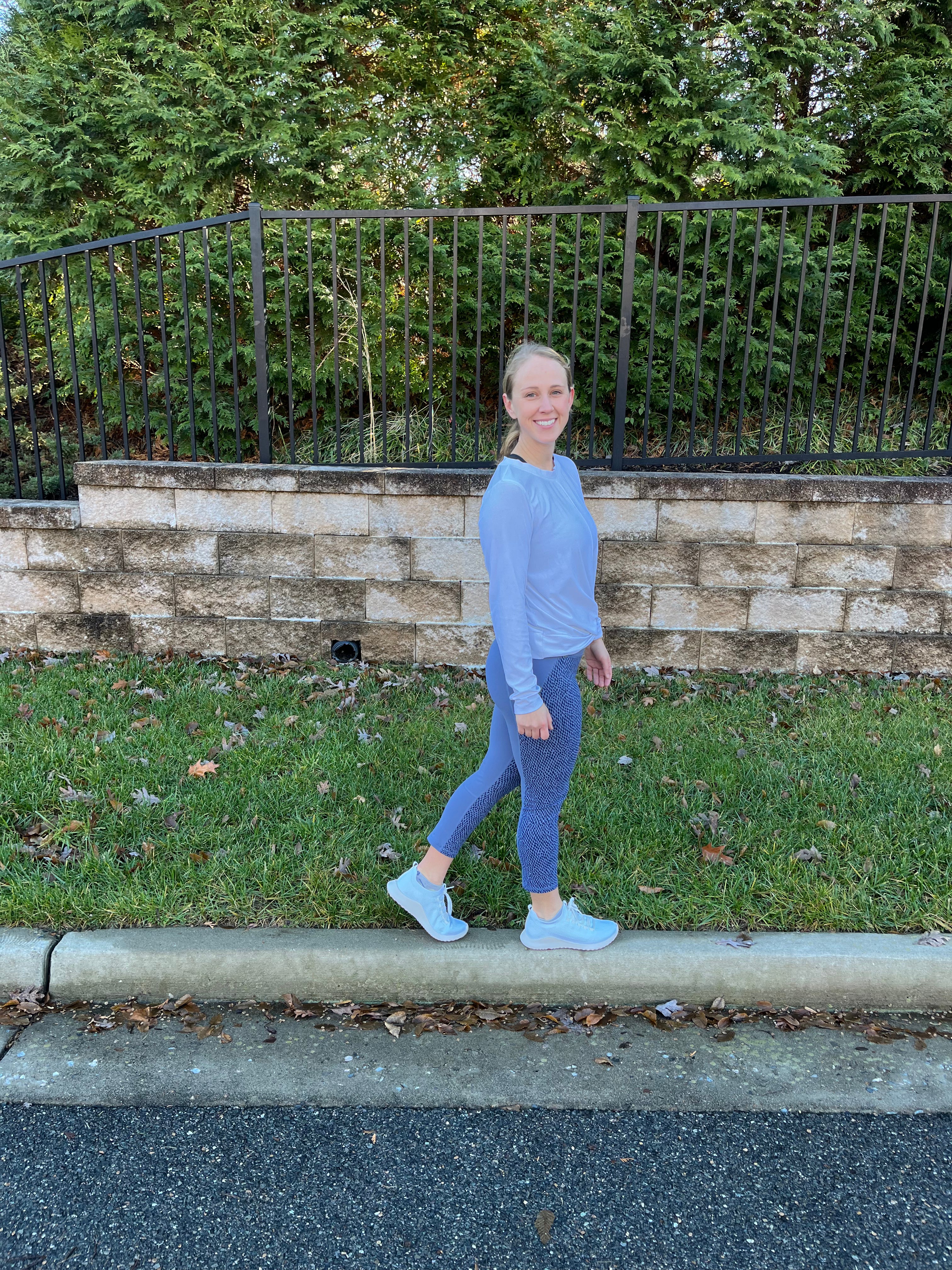 A woman wearing a matching light blue long-sleeve top and leggings set walks outdoors along a curb next to a stone wall, grass, and a black metal fence.