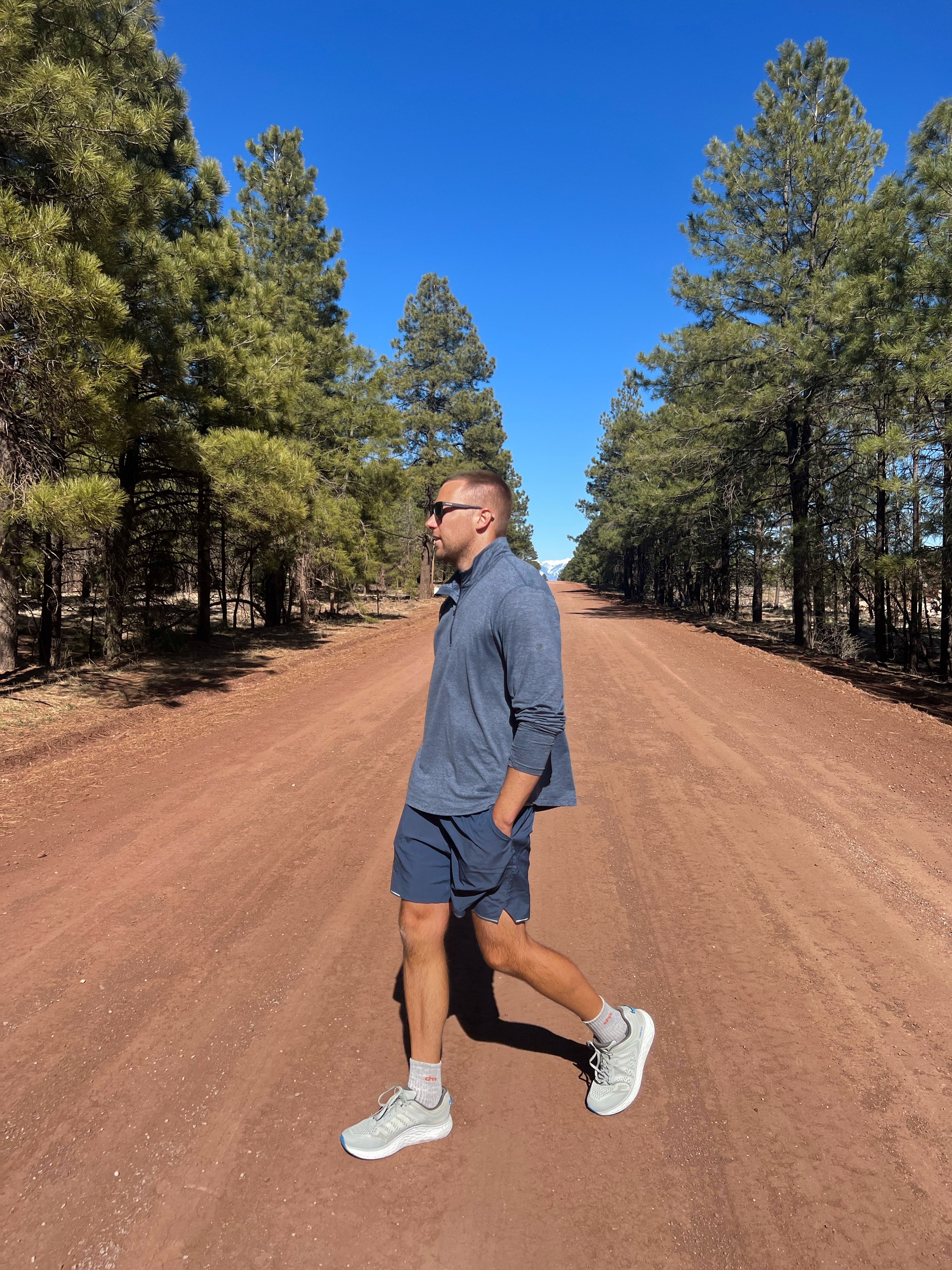 Man walking on a dirt road lined with tall trees under a clear blue sky.