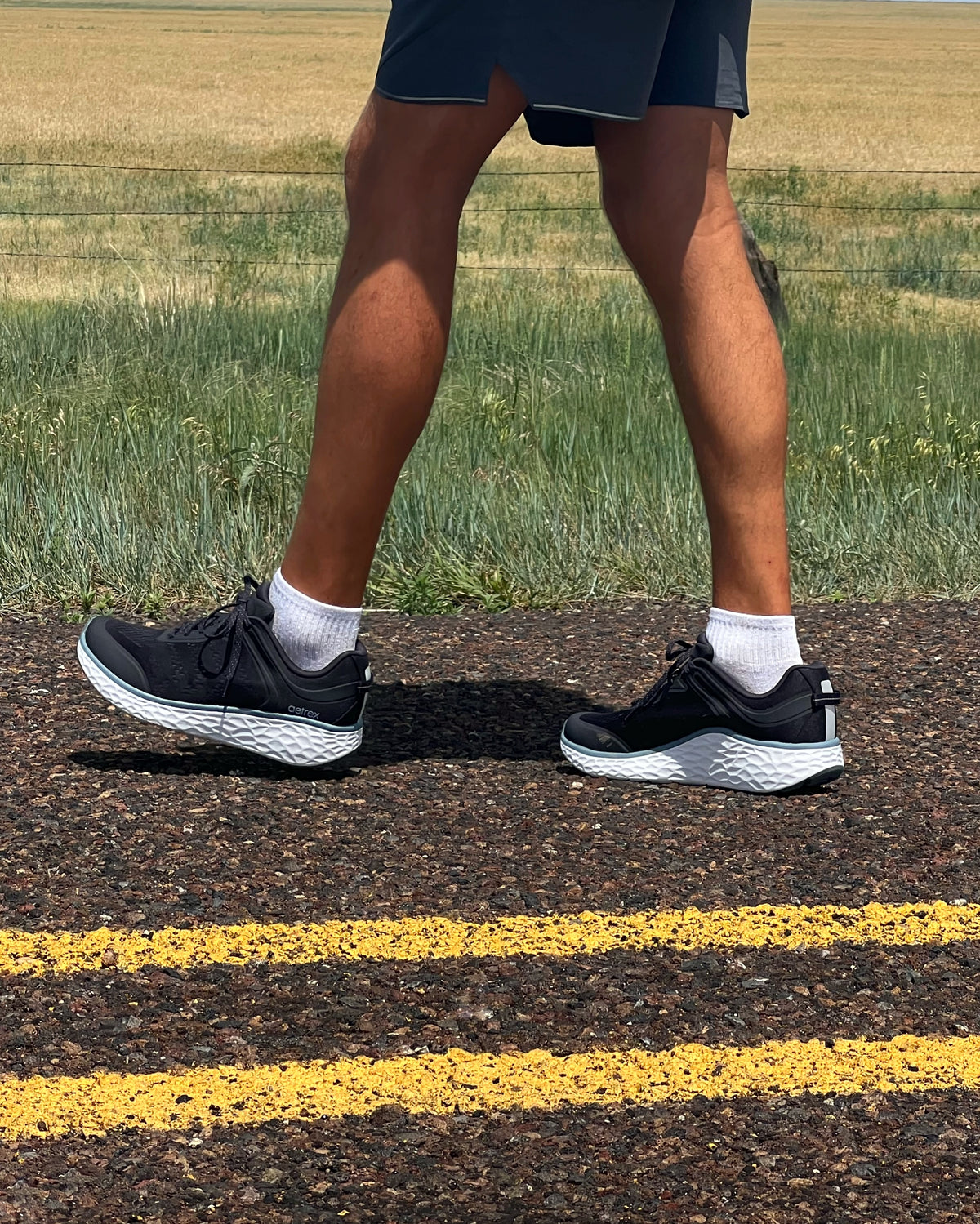 A person walking outdoors on a paved road wearing black athletic sneakers with white socks and dark shorts, with grass and a field in the background.