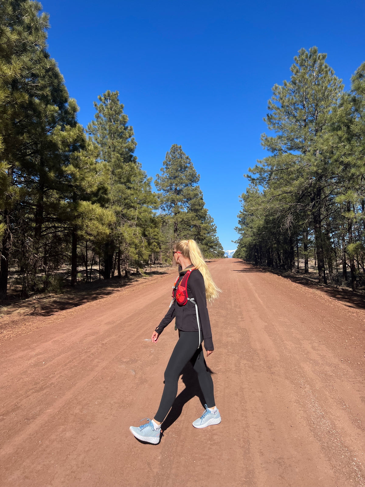 A woman walking on a dirt road between pine trees, wearing a black long-sleeve top, black leggings, light blue running shoes, and a red running backpack.