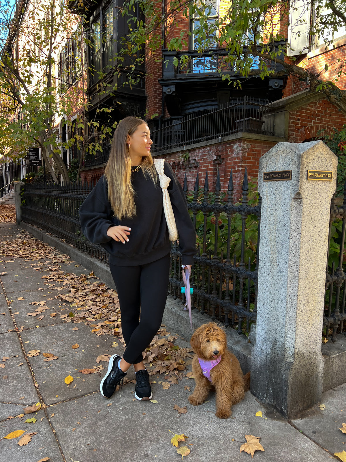 A woman in a black sweatshirt and black leggings stands on a city sidewalk next to a stone post, holding a pink umbrella while wearing a purple dog leash. A small brown dog wearing a purple bandana sits beside her.
