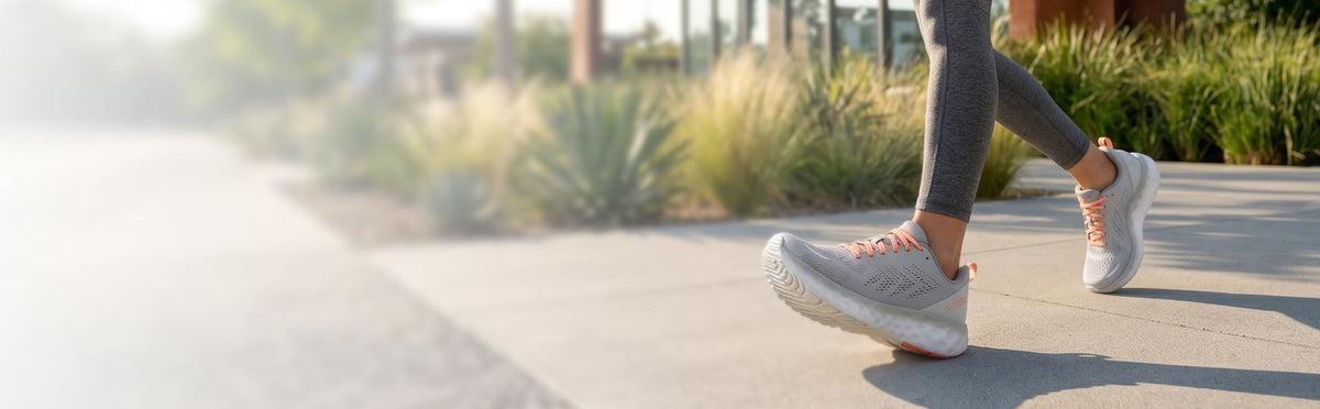 A person walking outdoors wearing light gray athletic sneakers with orange accents and gray leggings, photographed on a sunny paved path near modern landscaping and a glass building.