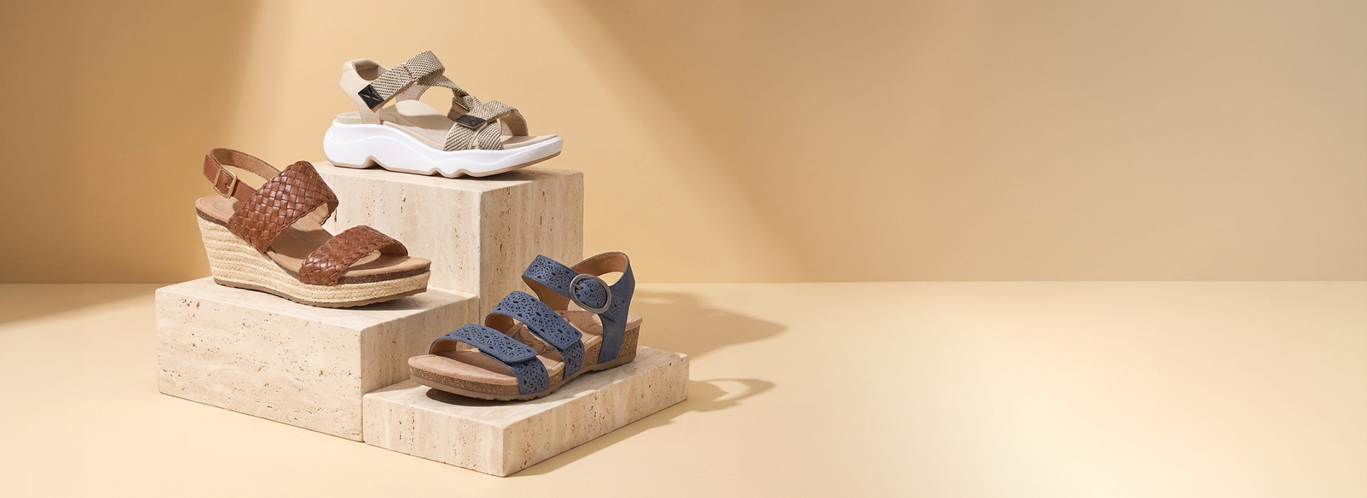 Three pairs of women's wedge sandals displayed on stacked stone blocks against a warm beige studio background. The sandals include brown leather, blue leather, and beige/white woven and fabric styles.