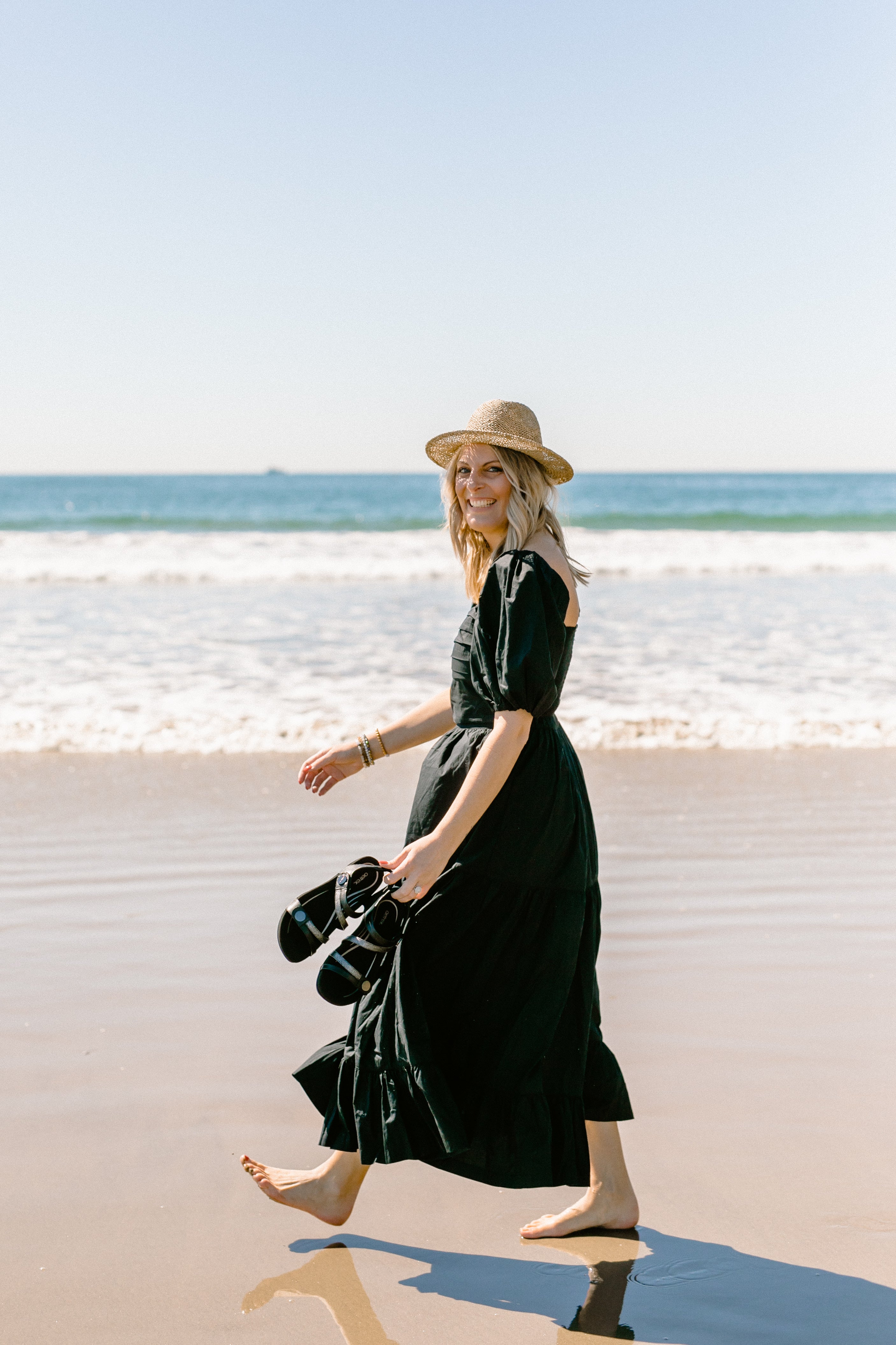 Woman in a black dress and straw hat standing on a beach with ocean waves in the background.