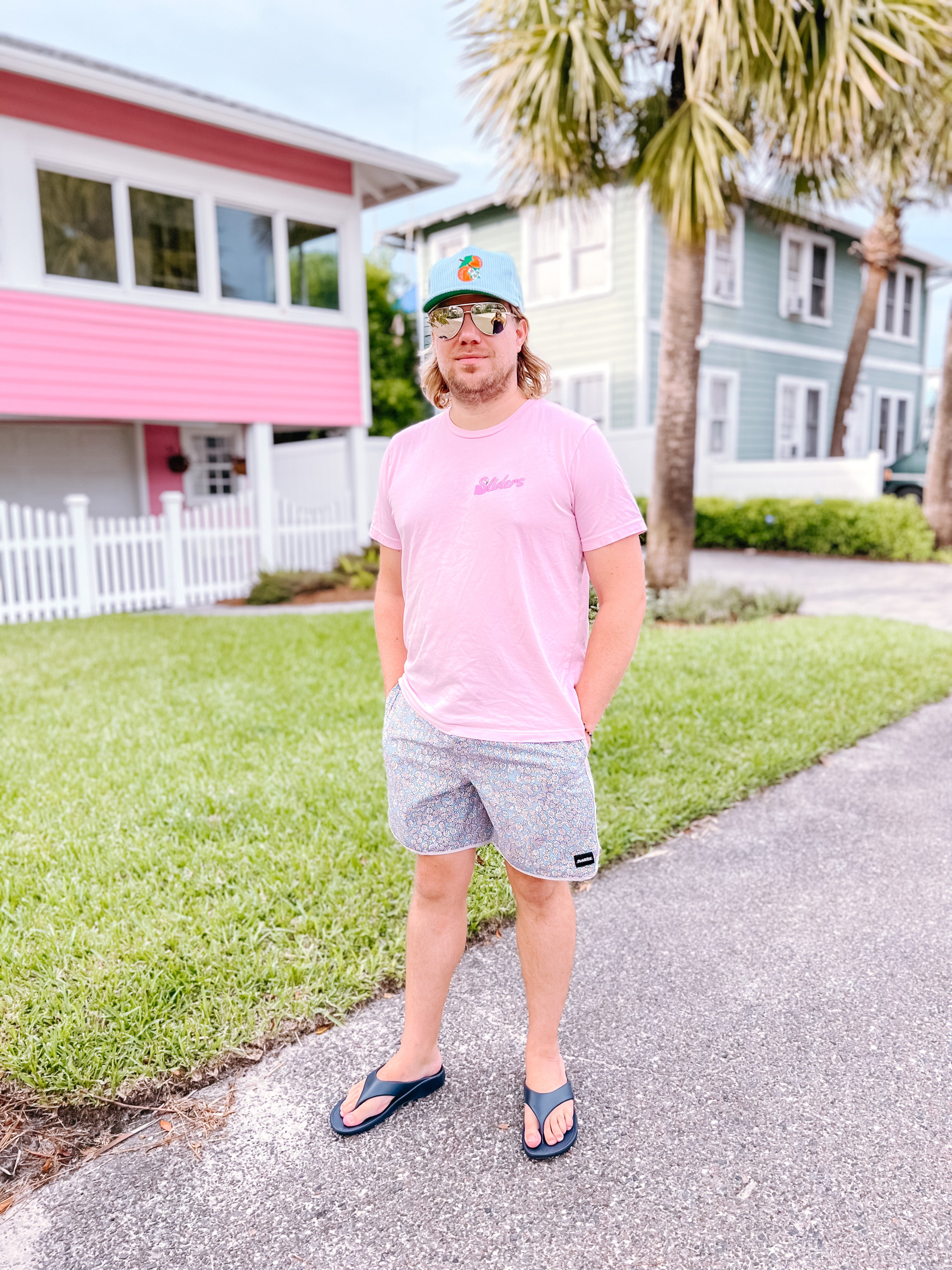 Man in pink t-shirt and gray shorts standing on a sidewalk with houses and palm trees in the background.