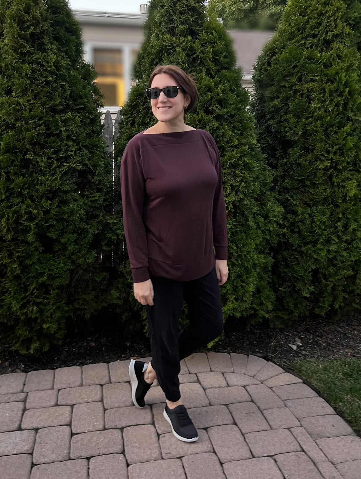 A woman wearing a dark plum long-sleeve top, black pants, and black sneakers stands on a stone path in front of tall green hedges.