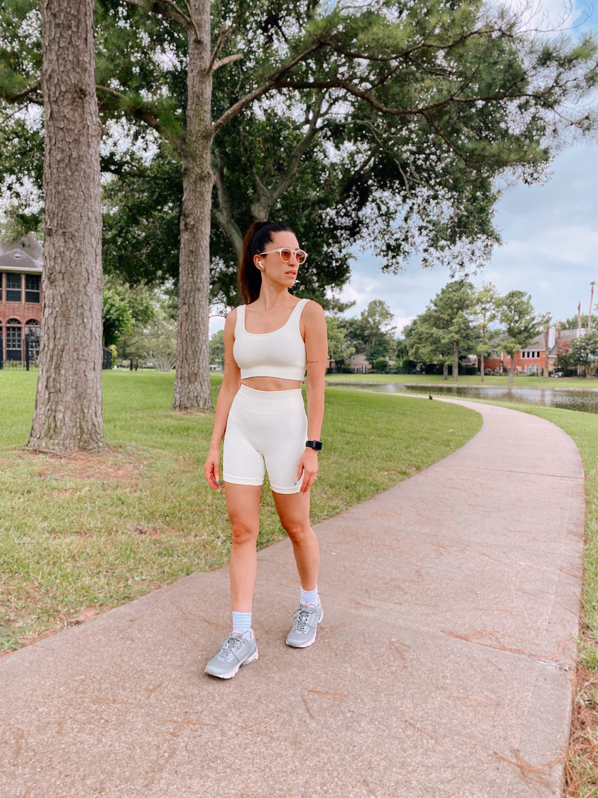 A woman wearing a matching cream-colored athletic set consisting of a tank top and shorts, standing on a paved path in a park with trees and grass.