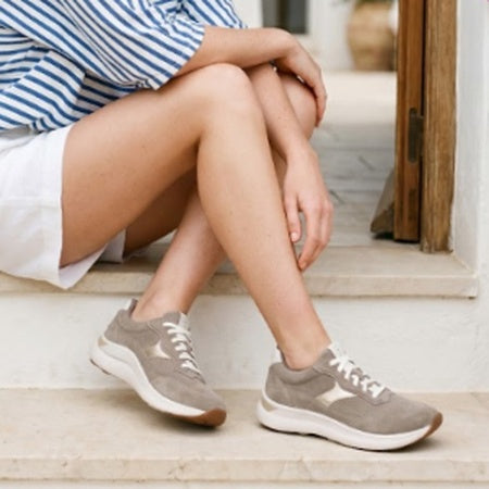 A person sitting on stone steps wearing taupe-brown and white low-top sneakers, with a blue-and-white striped shirt and white shorts visible.