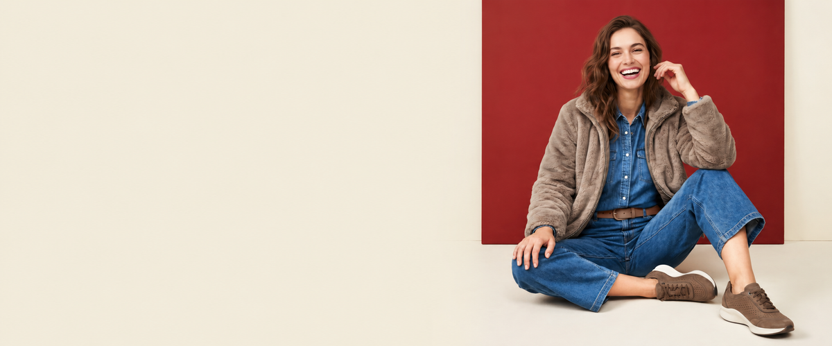A woman sitting cross-legged on a studio floor, smiling and posing in a brown fuzzy zip-up jacket, blue denim shirt and jeans, and brown sneakers. She is positioned against a split background of off-white on the left and a solid dark red on the right.