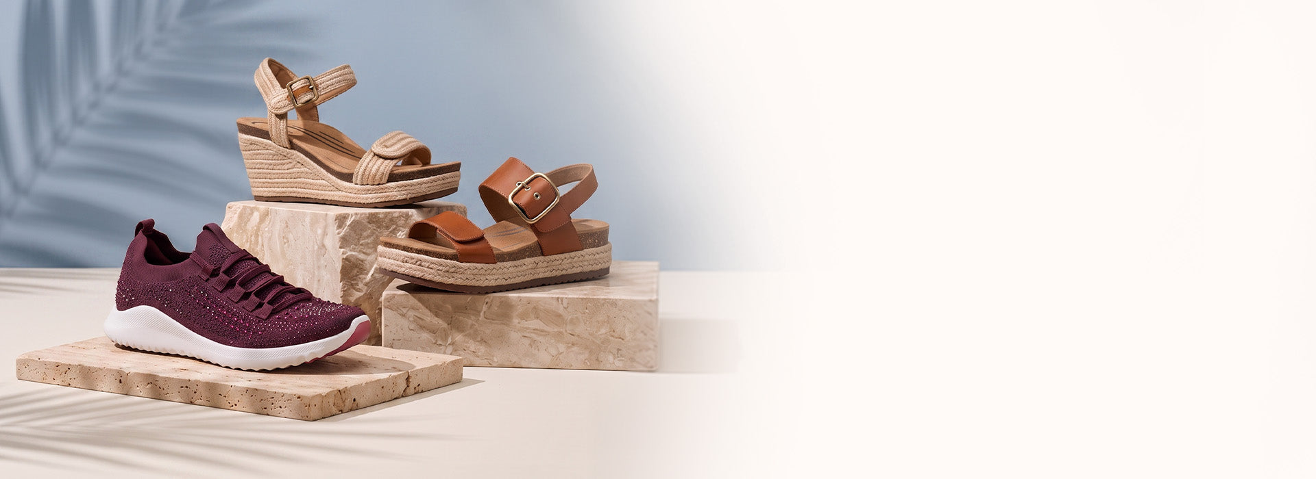 Three women's shoes displayed on stone blocks against a light blue backdrop with palm leaf shadows: a burgundy knit sneaker with a white sole, a tan wedge sandal, and a brown espadrille sandal with a buckle strap.