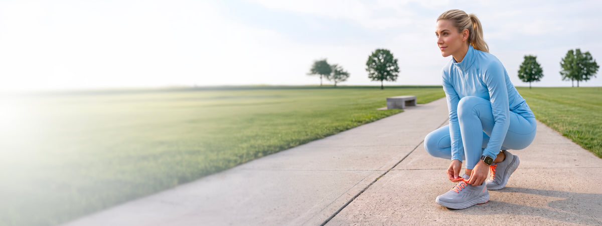 A woman in a light blue athletic tracksuit crouches on a concrete path in a park, tying the laces on her gray running shoes.