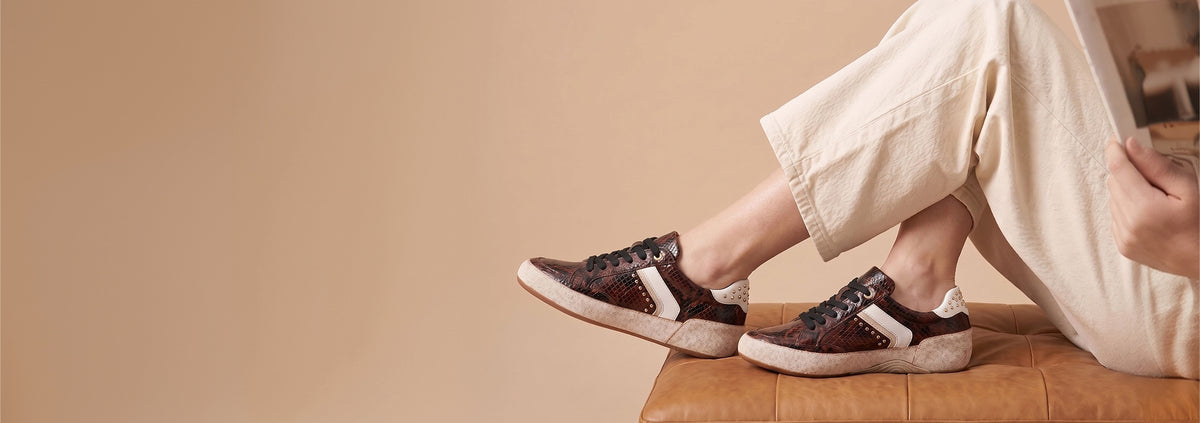 A person wearing brown snakeskin-patterned sneakers with beige side stripes and white soles, seated on a tan leather bench against a warm beige background.