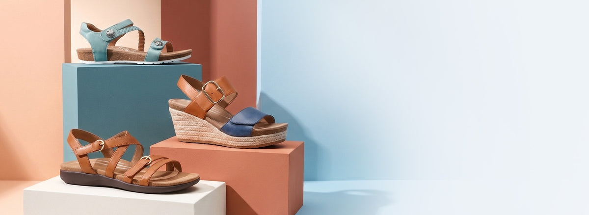 A styled product display of several wedge sandals arranged on geometric blocks against a split pastel background of peach and light blue.