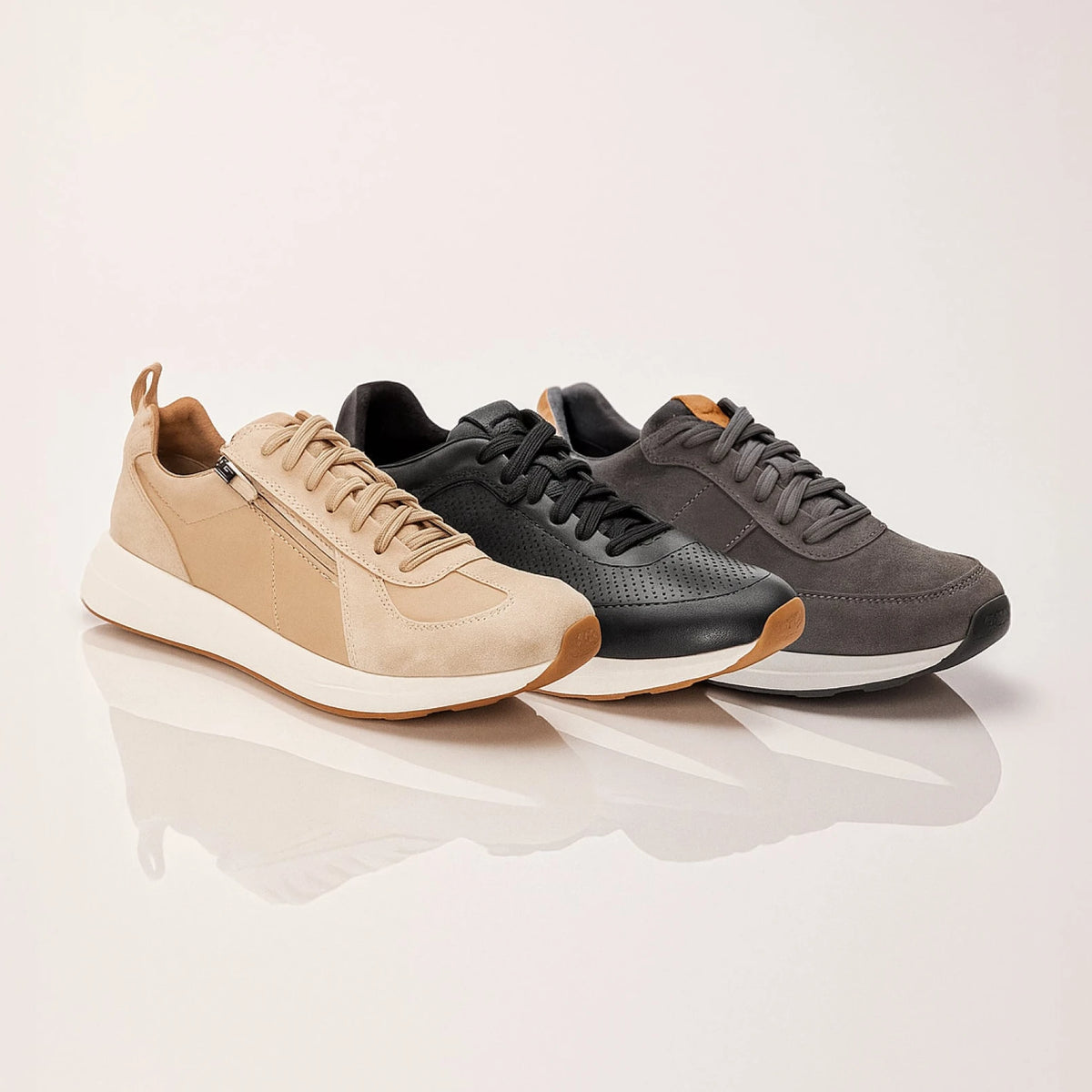 Three low-top sneakers arranged in a row on a light beige reflective surface: a tan sneaker in front, a black sneaker in the middle, and a dark gray sneaker behind. The shoes have lace-up fronts, side zippers on the tan and gray models, and thick white midsoles.