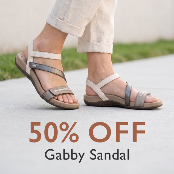 A person wearing a pair of flat strappy sandals with beige straps and gray accents, standing outdoors on a light concrete surface. Large text at the bottom reads '50% OFF Gabby Sandal'.