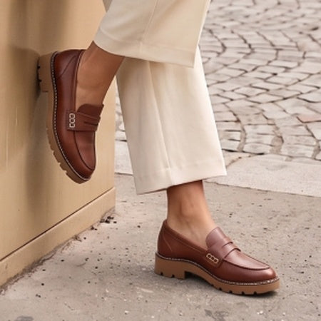 A person wearing brown leather penny loafers with beige wide-leg trousers, standing on a cobblestone street next to a beige wall.