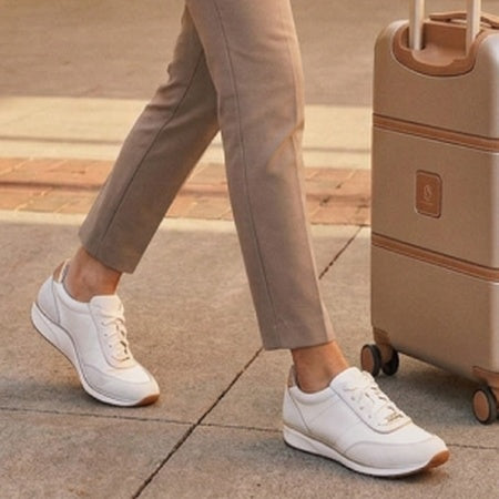 White low-top lace-up sneakers worn with beige trousers, shown on a person walking beside a beige rolling suitcase on a paved outdoor surface.