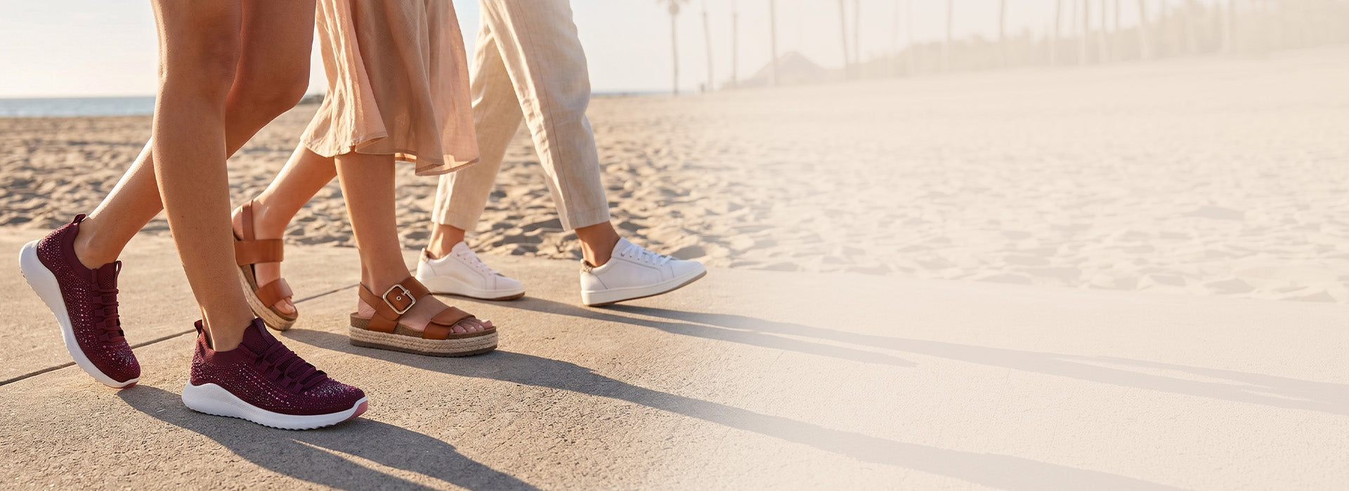 Three women walking along a sunny beachside path wearing blake sneakers, vania sandals, and carly sparkle sneaker with the ocean visible in the background.