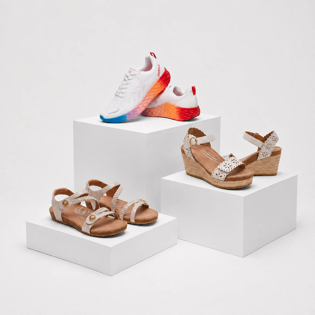 Three pairs of women's casual footwear displayed on white display blocks: a white sneaker with a red-to-blue gradient sole, tan wedge sandals with white lace-up straps, and tan flat sandals with wide straps and round gold-tone buttons.